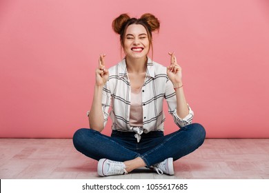 Portrait of a smiling young girl sitting on a floor with crossed legs and holding fingers for good luck isolated over pink background