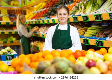 Portrait Of Smiling Young Girl Wearing Apron Standing In Fruit And Vegetable Store. First Job Concept..