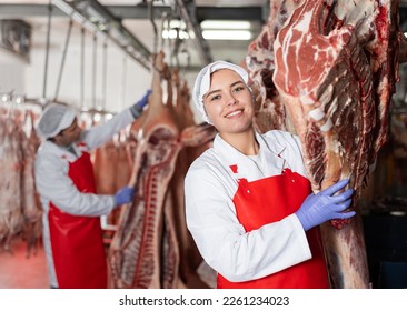 Portrait of smiling young girl in white coat and red apron working in butchery, standing in cold storage room near hanging raw cow carcasses.. - Powered by Shutterstock