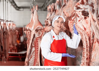 Portrait of smiling young girl in white coat and red apron working in butchery, standing in cold storage room near hanging raw cow carcasses.. - Powered by Shutterstock