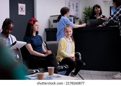 Portrait Of Smiling Young Girl With Worried Pensive Mother Sitting In Busy Clinic Reception With Diverse People. Parent With Child At Private Hospital Front Desk Waiting To See Pediatrician Specialist