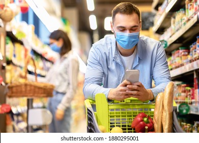 Portrait Of Smiling Young Guy In Medical Disposable Face Mask Doing Grocery Shopping, Using His Mobile Phone Gadget Device, Leaning On Trolley Cart. Male Consumer Looking For Sales On Internet Online