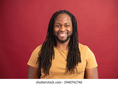 Portrait Of Smiling Young Handsome African American Guy With Dreadlocks In Yellow T-shirt Standing Isolated Over Red Background And Looking At The Camera, Cheerful Optimistic Real Person Black Man