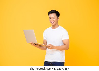 Portrait of a smiling  young handsome Asian man holding laptop computer while looking at camera in isolated studio yellow background