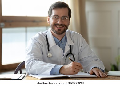 Portrait Of Smiling Young Handsome Male General Practitioner Therapist In Glasses And White Coat Sitting At Table In Hospital Office. Happy Physician Looking At Camera, Doing Paperwork At Workplace.