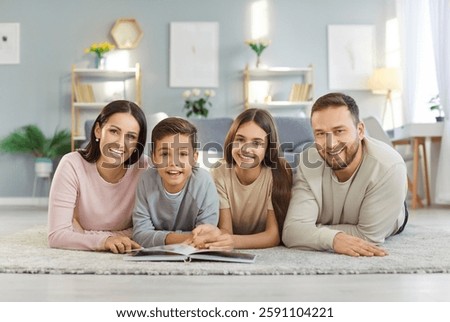Portrait of smiling young happy family of four with two children boy and girl lying on floor at home with photo album and looking cheerful at camera. Family time and leisure concept.