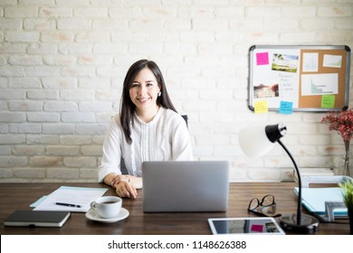 Portrait Of Smiling Young Hispanic Business Woman Sitting At Her Desk Table With A Laptop Computer Making An Eye Contact