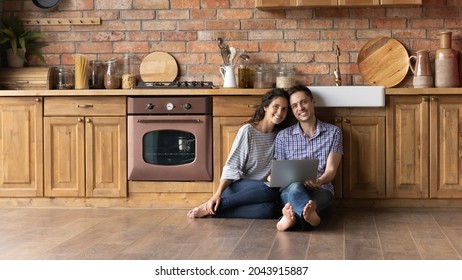 Portrait Of Smiling Young Hispanic Family Couple Sitting On Heated Warm Floor In Wooden Kitchen With Computer, Spending Free Weekend Leisure Time Online Shopping Or Web Surfing, Posing In Own House.