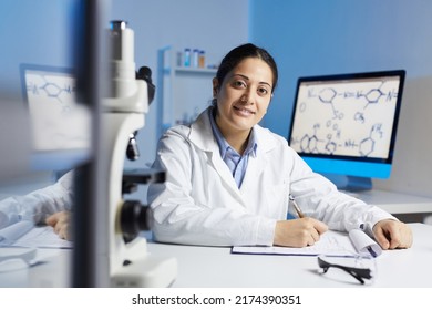 Portrait Of Smiling Young Indian Woman In Lab Coat Sitting At Desk And Making Notes In Clipboard While Working In Scientific Laboratory