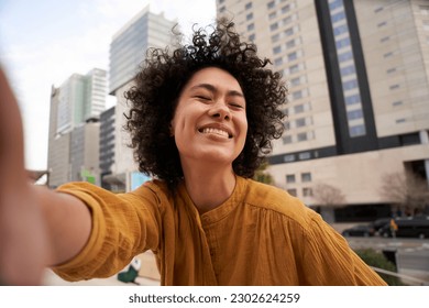 Portrait of smiling young Latin girl with closed eyes and afro hair taking a selfie outside a shopping mall. Happy millennial people using mobile phones and posing for photos social media.