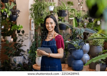 Portrait of smiling young latin woman florist standing and holding digital tablet at floral shop. Successful flower shop owner standing in plant store looking at camera. Happy mixed race entrepreneur.