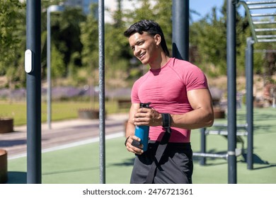 portrait of a Smiling young Latino man in pink shirt taking a break at an outdoor gym, holding a water bottle and enjoying a sunny day in the park. - Powered by Shutterstock