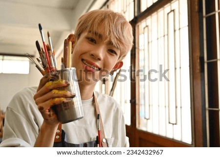 Portrait of Smiling Young LGBT teenage boy with colored hair holding paint brushes and equipment for drawing and painting, modern artists with art.