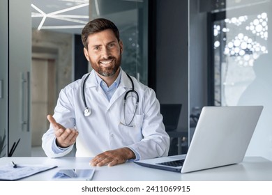 Portrait of a smiling young male doctor talking and consulting on a video call. He is sitting at the table in the office of the clinic, gesturing with his hand in front of the camera. - Powered by Shutterstock