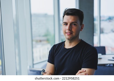 Portrait Of A Smiling Young Man In A Black T-shirt Against The Background Of A Modern Office. Stylish Guy In Casual Clothes