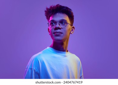 Portrait of smiling young man, boy in casual white t-shirt and sunglasses looking away against purple background in neon light. Relaxed look. Concept of human emotions, youth, education, lifestyle, ad