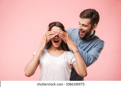 Portrait Of A Smiling Young Man Covering His Girlfriends Eyes As A Surprise Isolated Over Pink Background