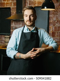 Portrait Of Smiling Young Man With Facial Hair Wearing Apron And Leaning Against Bar Counter In Cafe. Blank Slate Board Copy Space For Text Or Image.Cozy Coffee Shop With Bartender Owner Staff Service