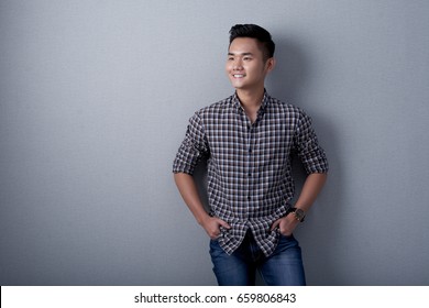 Portrait Of Smiling Young Man With Hands In Pockets Leaning On Gray Wall And Looking Away Dreamily, Studio Shot