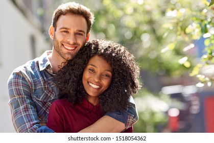 Porträt eines lächelnden jungen Mannes, der seine Freundin von hinten umarmt, während er an einem sonnigen Tag zusammen in der Stadt steht
