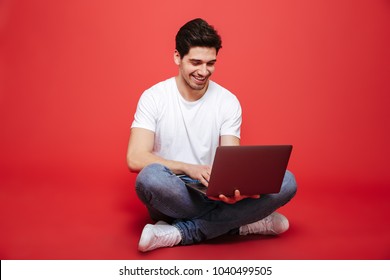 Portrait of a smiling young man in white t-shirt using laptop computer while sitting on a floor isolated over red background