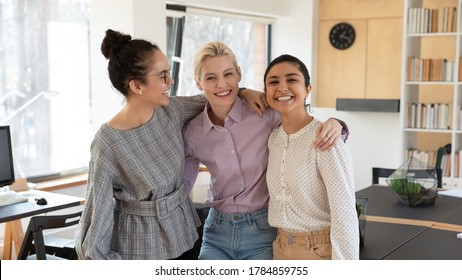 Portrait of smiling young multiracial female colleagues hug posing in modern office together, happy millennial diverse multiethnic women employees embrace at workplace, show friendship and unity