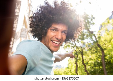 Portrait Of Smiling Young North African Man With Afro Hair Taking Selfie Outside In City And Pointing 