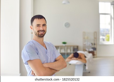 Portrait of smiling young professional masseur or physiotherapist looking at camera standing arms folded against blurred background of massage room in modern health center