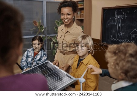 Portrait of smiling young teacher showing solar panel to class of little kids learning about renewable energy