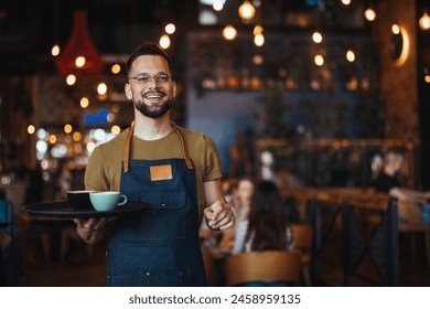 Portrait of smiling young waiter holding tray in cafe. Handsome waiter smiling at camera holding tray at the coffee shop. Profesional Waiter in Restaurant  - Powered by Shutterstock