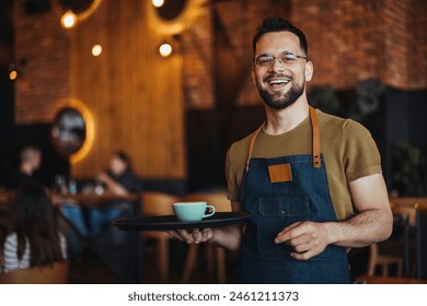 Portrait of smiling young waiter holding tray in cafe. Handsome waiter smiling at camera holding tray at the coffee shop. Profesional Waiter in Restaurant 