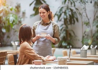 Portrait of smiling young waitress bringing coffee to client at outdoor cafe terrace lit by warm sunlight, copy space - Powered by Shutterstock