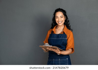 Portrait of smiling young woman in apron looking at camera. Beautiful latin waitress isolated on grey wall. Successful small business owner standing on grey background with copy space. - Shutterstock ID 2147820679
