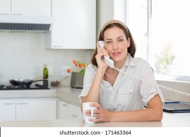 Portrait Of A Smiling Young Woman With Coffee Cup Using Landline Phone In The Kitchen At Home