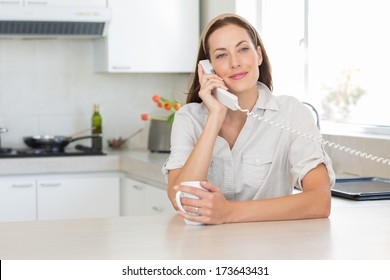 Portrait Of A Smiling Young Woman With Coffee Cup Using Landline Phone In The Kitchen At Home