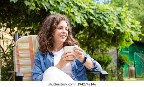 Portrait Of Smiling Young Woman Enjoying Drinking Coffee Or Tea Sitting On Chair On Home Terrace On Green Garden Background. Happy Morning Routine, Day Planning, Enjoying Life, Here And Now Concept