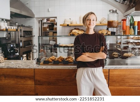 Portrait of smiling young woman entrepreneur standing at the counter of her bakery and coffee shop. Local small business owner indoors. Female in cafe near showcase with fresh croissants and bread.