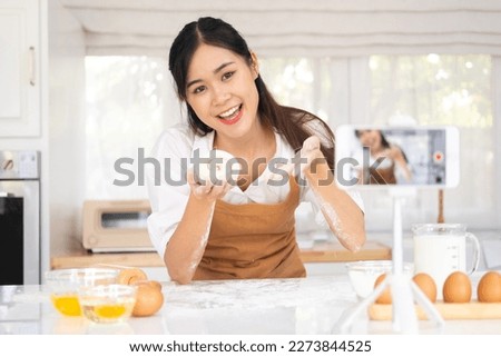 Portrait of smiling young woman food vlogger showing process of baking in live stream, sitting in front of smart phone on tripod, broadcasting. Indoor studio shot on kitchen background.