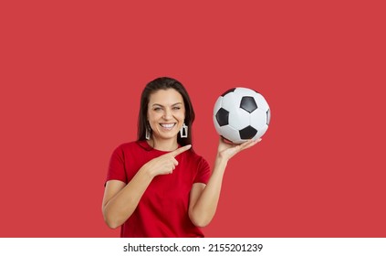 Portrait Of Smiling Young Woman Isolated On Red Studio Background Hold Point At Ball Cheering Football Team. Happy Female Fan Enjoy Sports Activity Support Players. Hobby Concept.