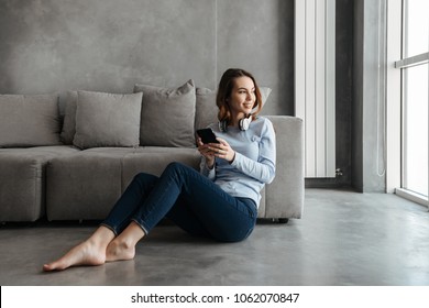 Portrait Of A Smiling Young Woman Listening To Music With Headphones And Using Mobile Phone While Leaning On A Couch At Home