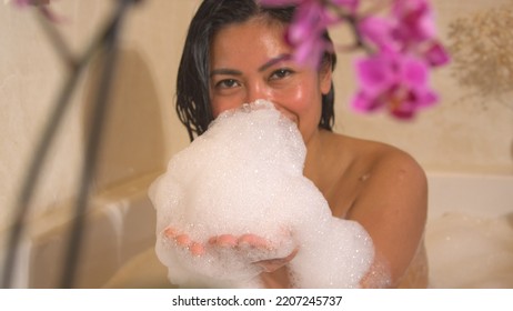 PORTRAIT: Smiling Young Woman Looking Into Camera With Hands Full Of Bath Foam. Female Person Laughing And Enjoying In Bathtub Full Of Bubble Foam. Home Relaxation In Bubbly Bath After A Long Busy Day