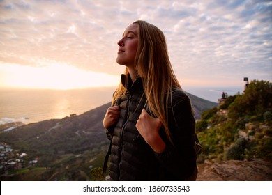 Portrait of smiling young woman on vacation closing her eyes as she walks along coastal path and sun sets over sea behind her - Powered by Shutterstock