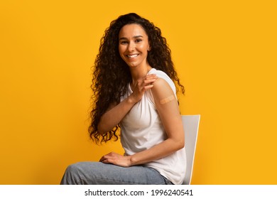 Portrait Of Smiling Young Woman Showing Arm After Coronavirus Vaccination, Happy Brunette Female Had Covid Vaccine Injection, Sitting On Chair With Rolled Up Sleeve Over Yellow Background, Copy Space