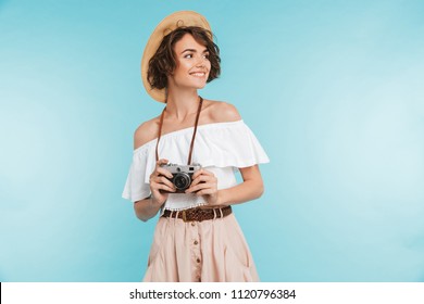 Portrait of a smiling young woman in summer hat standing with photo camera and looking away at copy space isolated over blue background - Shutterstock ID 1120796384