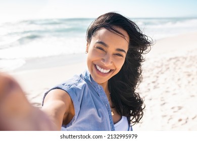 Portrait of smiling young woman taking a selfie at beach during. Cheerful hispanic woman enjoying at beach during holiday. Happy girl taking photo over exotic tropical beach looking at camera. - Powered by Shutterstock