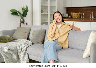Portrait of smiling young woman talking by smartphone and looks away. Carefree charming asian lady enjoys phone conversation with friend, sits on sofa, chatting and laughing