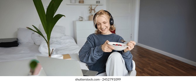 Portrait of smiling young woman, watching tv show in headphones, eating breakfast and looking at laptop screen. - Powered by Shutterstock