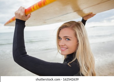 Portrait Of A Smiling Young Woman In Wet Suit Holding Surfboard Over Head At Beach