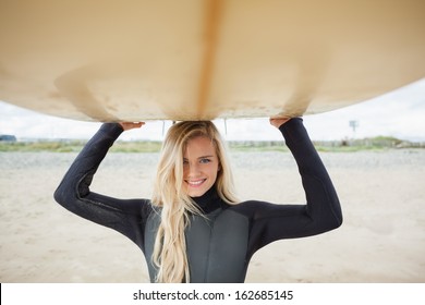 Portrait Of A Smiling Young Woman In Wet Suit Holding Surfboard Over Head At Beach