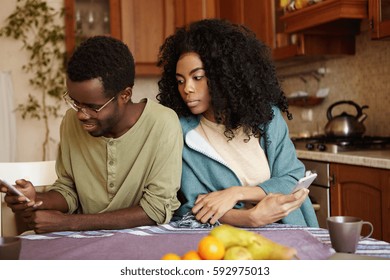 Portrait Of Sneaky And Jealous Young African Wife Spying On Her Husband, Looking Over Shoulder, Trying To See What He Reading On His Mobile Phone. People, Relationships And Modern Technology